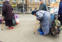 Сгорбленная старушка стоит, облокотившись о столб. Сумка рядом. Парни кладут туда еду. //Фото Инна Лебедева